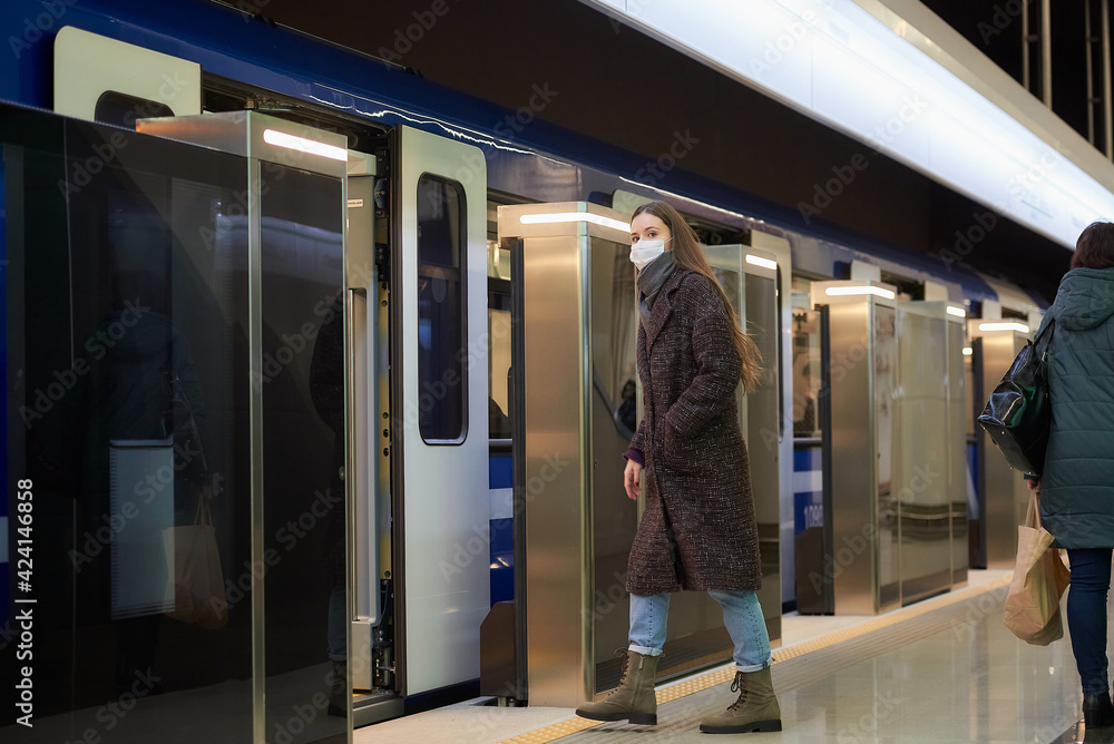 A full-length photo of a woman in a medical face mask to avoid the spread of coronavirus who is entering the modern subway car. A girl in a surgical mask is keeping social distance on a metro station. A full-length photo of a woman in a medical face mask to avoid the spread of coronavirus who is entering the modern subway car. A girl in a surgical mask is keeping social distance on a metro station.
