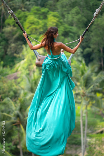 Tanned young woman riding on a long swing in long turquoise dress. Island of Bali. Tropical forest on the background. Travel and joy. Close up