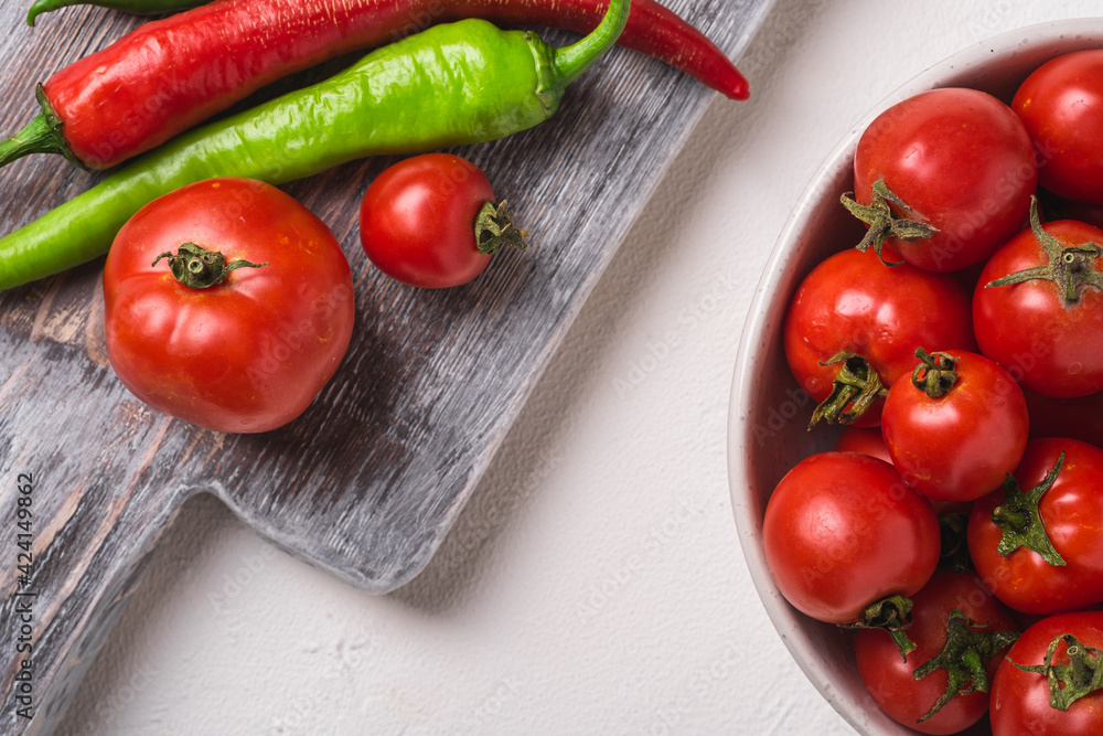 Fresh ripe tomatoes in bowl near to old wooden cutting board with hot chili peppers, white stone concrete background, top view macro Fresh ripe tomatoes in bowl near to old wooden cutting board with hot chili peppers, white stone concrete background, top view macro