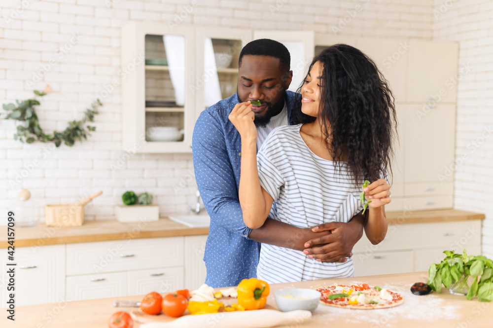 Lovely African American couple standing next to kitchen counter, hugging, spending leisure time weekend together at home, girlfriend looking at boyfriend and giving to smell herbs mint, cooking pizza