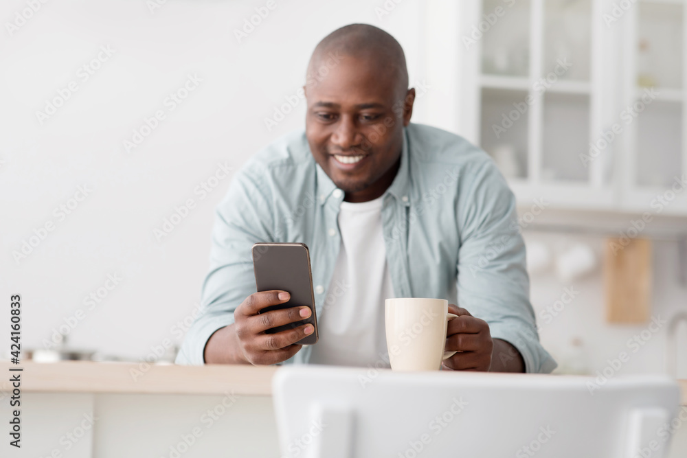 Happy black mature man drinking morning coffee and using smartphone in kitchen, enjoying hot drink at home Happy black mature man drinking morning coffee and using smartphone in kitchen, enjoying hot drink at home