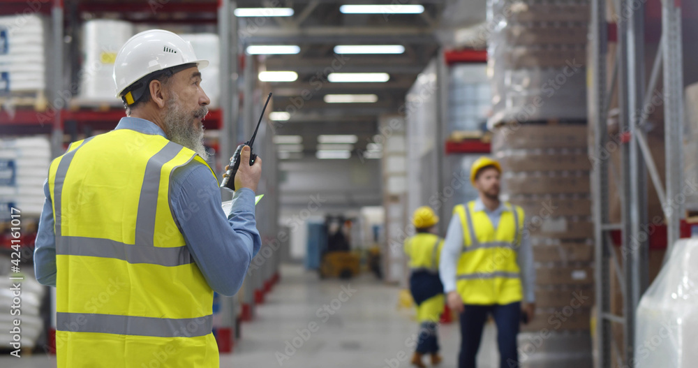 Senior manager in safety uniform transferring message to employee with walkie-talkie in warehouse Senior manager in safety uniform transferring message to employee with walkie-talkie in warehouse
