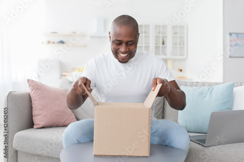 Happy black man unpacking delivery box, sitting on couch at home interior. Satisfied customer opening package