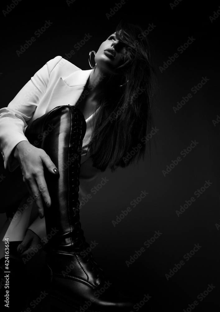 Obraz premium Black and white. Gorgeous tall sexy woman in white jacket stands holding foot up, step on, touches her high leather boot and looks up. Fashion, vogue, sexy stylish look for woman concept