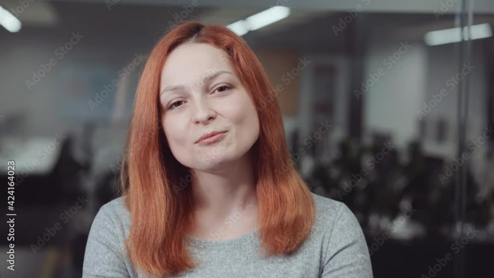 Red-haired woman looking at camera and talking, blurred office on background. Online business meeting or job interview with participants using webcams. Concept of distance communication Red-haired woman looking at camera and talking, blurred office on background. Online business meeting or job interview with participants using webcams. Concept of distance communication