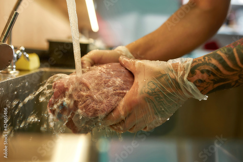 Close up shot of hands of male cook wearing protective gloves rinsing raw pork meat under tap water for cooking