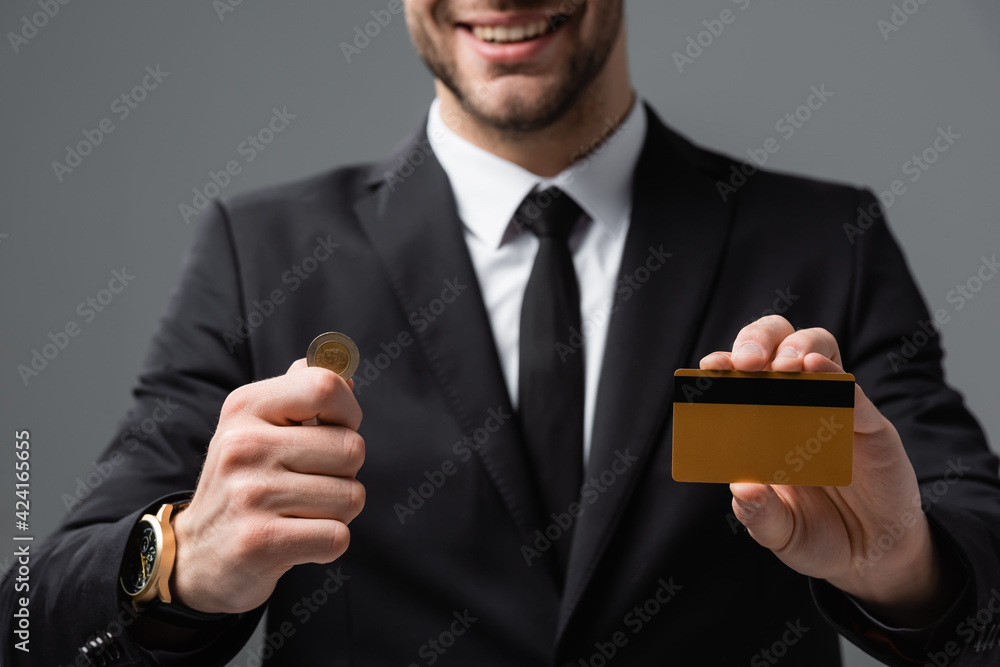 cropped view of smiling businessman holding coin and credit card on blurred background isolated on grey cropped view of smiling businessman holding coin and credit card on blurred background isolated on grey