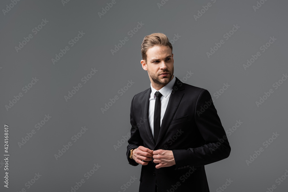 young businessman buttoning blazer while looking away isolated on grey young businessman buttoning blazer while looking away isolated on grey