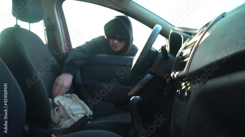 a man in a black hoodie and black glasses steals a bag from the seat of someone else's car. transportation, crime and ownership concept