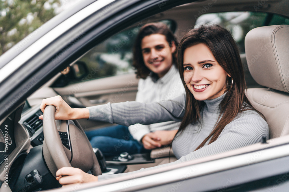 Happy smiling girl behind the wheel of a modern car looks into the camera, next to her in the front seat of her young boyfriend, enjoying the journey