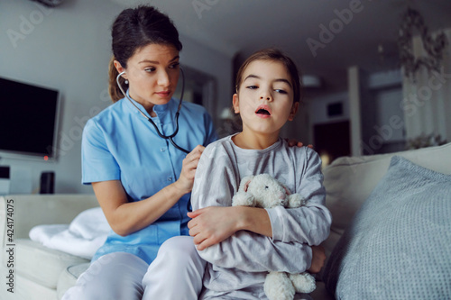 Doctor sitting on sofa next to girl and examining her lungs with stethoscope. Doctor at home service.