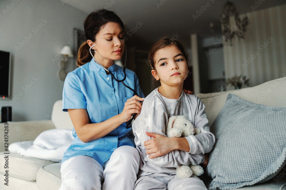 Doctor sitting on sofa next to girl and examining her lungs with stethoscope. Doctor at home service. Doctor sitting on sofa next to girl and examining her lungs with stethoscope. Doctor at home service.