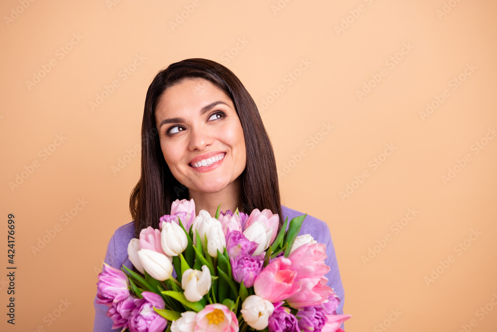 Photo of nice optimistic brunette long hairdo lady hold flowers look empty space wear purple sweater isolated on pastel beige color background Photo of nice optimistic brunette long hairdo lady hold flowers look empty space wear purple sweater isolated on pastel beige color background