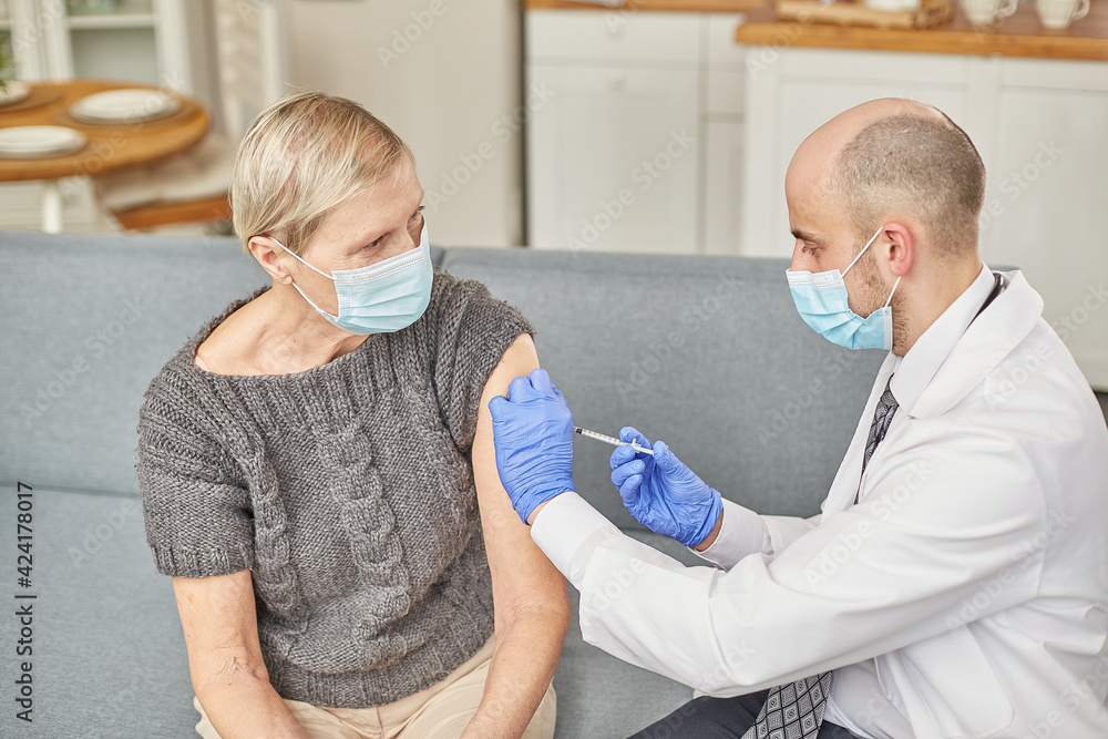 Fototapeta premium Man doctor vaccinates an elderly woman at her home wearing a protective mask. Vaccination concept