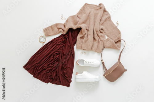 Female spring look autumn outfit burgundy skirt beige sweater white shoes sneakers bag. Folded fly clothes for women fashion urban basic outfit with accessories. Top view on white background. Flat lay photo