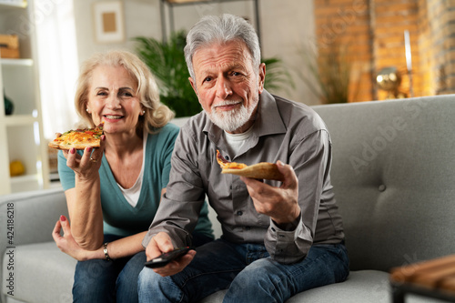  Cheerful husband and wife sitting on sofa at home. Happy senior woman and man eating pizza while watching a movie