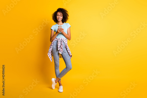Full length photo of sweet dark skin wavy lady wear blue t-shirt holding modern gadget empty space isolated yellow color background © deagreez