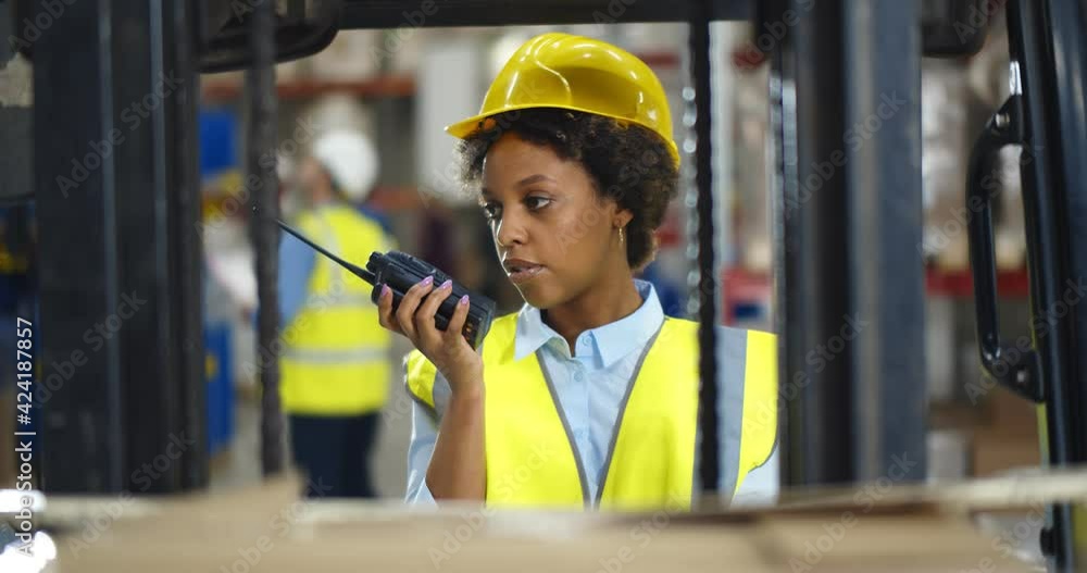 Young african woman standing near forklift and using walkie-talkie in warehouse Young african woman standing near forklift and using walkie-talkie in warehouse