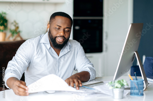 Busy successful African American man, entrepreneur or lawyer, in formal wear, sits at work desk, uses computer for work, studies financial documents, analysis profit and strategy of company