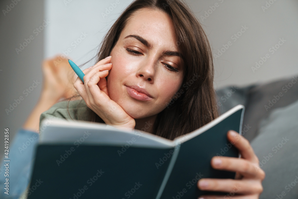 Fototapeta premium Young white woman reading her planner while lying on sofa at home