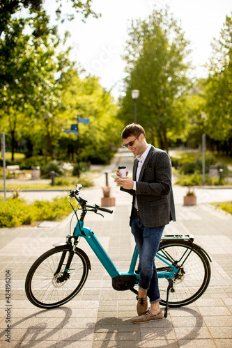 Young businessman using mobile phone by the ebike with takeaway coffee cup