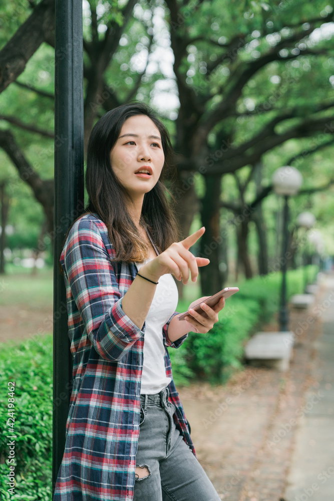 Naklejka premium charming young asian japanese woman standing by light pole in park pathway reading online map app information on mobile phone. cute girl holding cellphone point aside searching right way in garden.