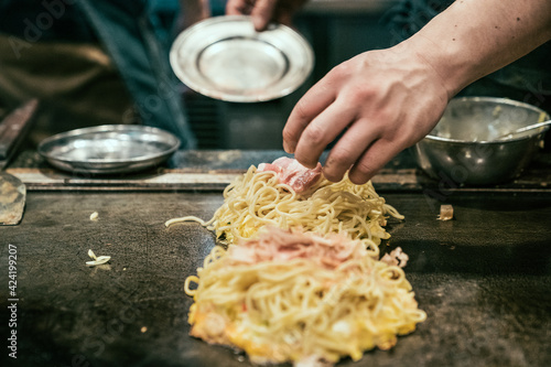 okonomiyaki Japanese pizza on hot metal pan. male chef hands adding meat on top noodles and eggs while cooking in teppanyaki place. local specialty food in osaka delicious okonomiyaki on iron plate photo