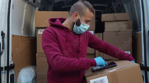 Portrait of a young man shipper with his van during the delivery round in the global pandemic from Covid-19 Coronavirus wearing face mask while checking address with tablet