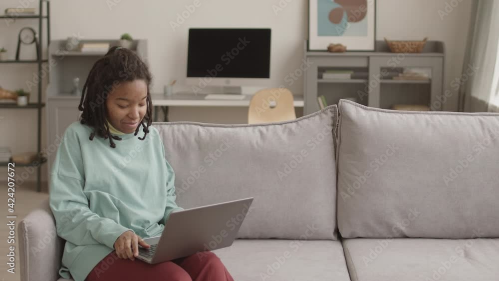 Medium shot of young African-American woman with problem skin and dreads on her head sitting on couch in living room and having video conversation using laptop Medium shot of young African-American woman with problem skin and dreads on her head sitting on couch in living room and having video conversation using laptop