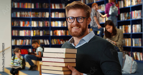 Bearded school teacher or librarian holding books in hands standing in library interior. photo