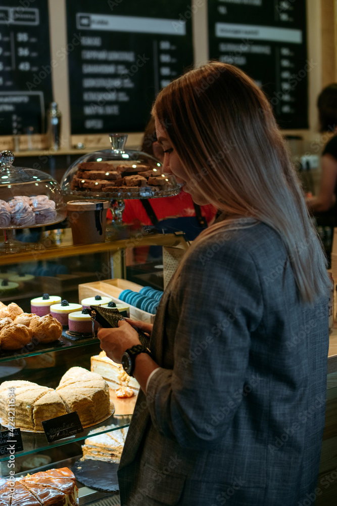 Confectionery customer, client in sweet-shop, pastry-shop. Back view of young woman choosing cake desserts from display in confectionery, shop, cafe.