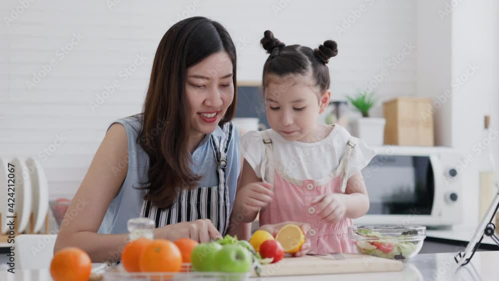 Family single mother and daughter enjoy and happy cooking in clean kitchen. Parents help children make fruit and vegetable salads from Video tutorial from tablet. Learn cook homemade from video call. Family single mother and daughter enjoy and happy cooking in clean kitchen. Parents help children make fruit and vegetable salads from Video tutorial from tablet. Learn cook homemade from video call.