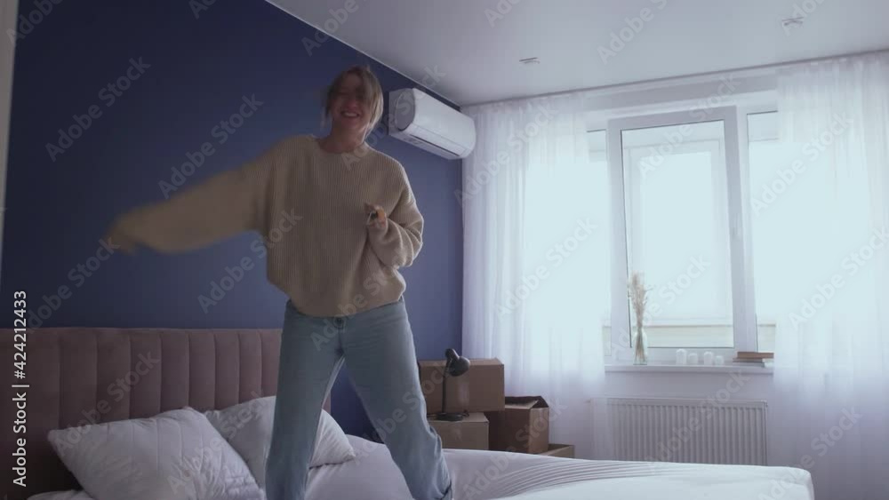 Happy young woman wearing jeans and sweater dances jumps on bed at home Happy young woman wearing jeans and sweater dances jumps on bed at home