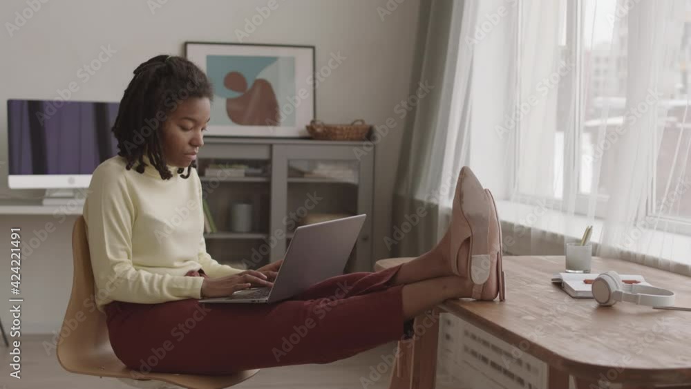 Lockdown of young African-American woman with dreads wearing casual clothes sitting on chair with laptop on her knees and legs on desk and working Lockdown of young African-American woman with dreads wearing casual clothes sitting on chair with laptop on her knees and legs on desk and working