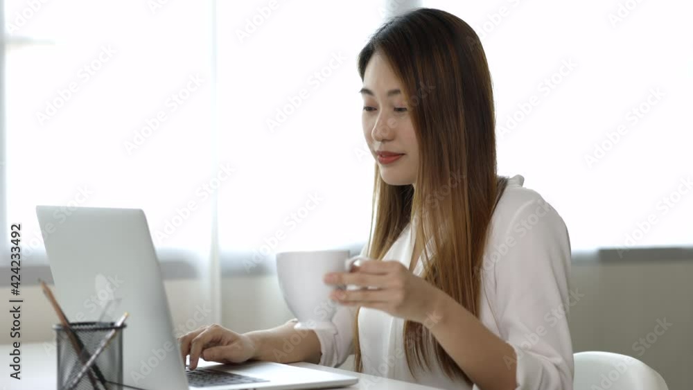 Young asian beautiful business woman working with laptop sitting at home drinking coffee. Smiling charming happy young female doing homework meeting conference with team at home. Young asian beautiful business woman working with laptop sitting at home drinking coffee. Smiling charming happy young female doing homework meeting conference with team at home.