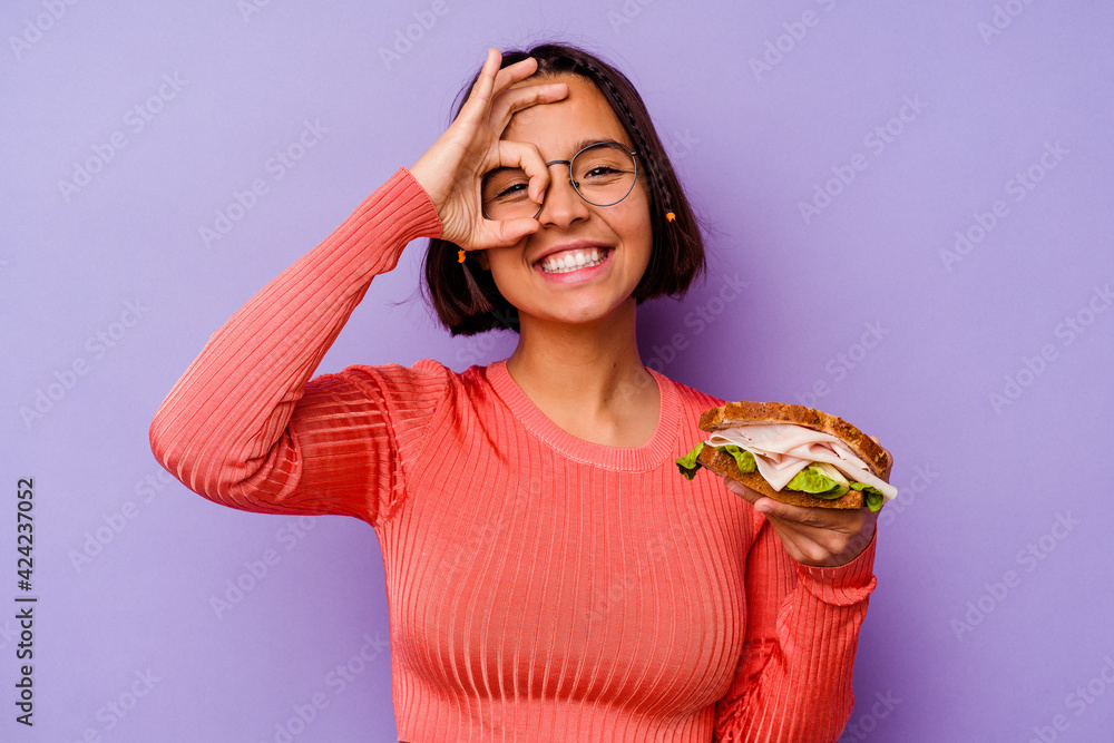 Young mixed race woman holding a sandwich isolated on purple background excited keeping ok gesture on eye. Young mixed race woman holding a sandwich isolated on purple background excited keeping ok gesture on eye.