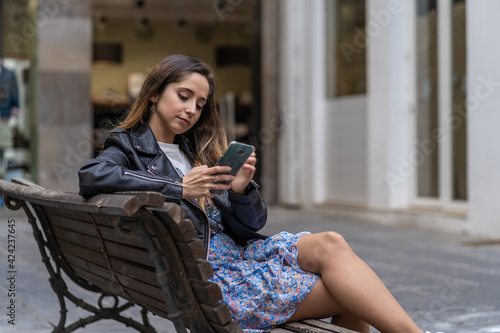 woman sitting on a bench with a smartphone checking her messages. With skirt and leather jacket