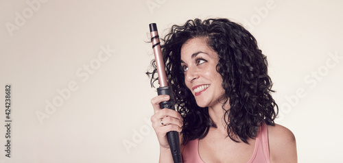 Young smiling woman with black curly hair uses tweezers or curling iron to get curls and waves in her hair. care and beauty concept. copy space.