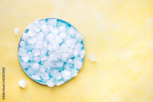Heap Of Ice Cubes And Crushed Ice On Blue Plate From Above. Yellow  Background. Blank space