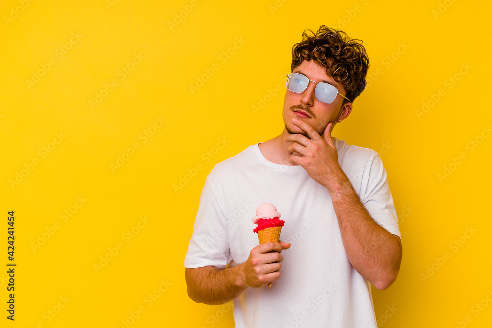 Young caucasian man eating an ice cream isolated on yellow background looking sideways with doubtful and skeptical expression. Young caucasian man eating an ice cream isolated on yellow background looking sideways with doubtful and skeptical expression.