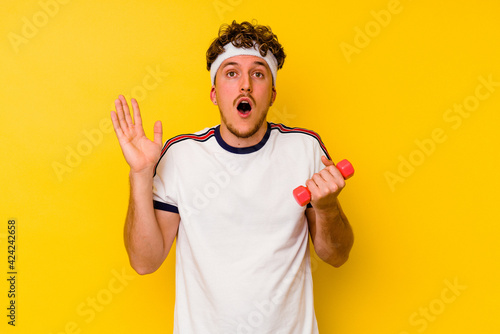 Young sport caucasian man holding a dumbbell isolated on a yellow background surprised and shocked.