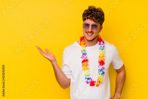 Young caucasian man wearing a Hawaiian party stuff isolated on yellow background showing a copy space on a palm and holding another hand on waist.