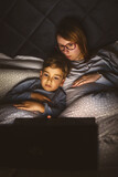 Mother and son watching movie or cartoon using laptop in bed at nighttime - Small boy and his mom using computer while lying in bed at night in dark room - leisure activity family bedtime concept