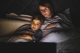 Mother and son watching movie or cartoon using laptop in bed at nighttime - Small boy and his mom using computer while lying in bed at night in dark room - leisure activity family bedtime concept