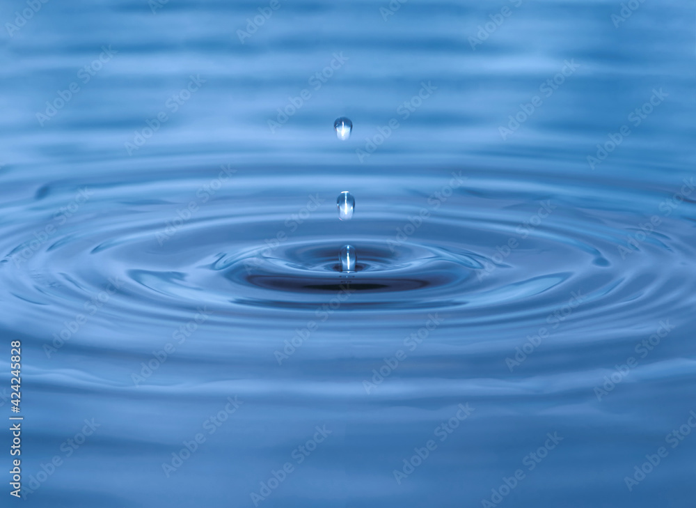 Water droplet dripping into blue pool creating ripples and abstract shape of water. Darkest area in center is in camera focus. Image contains some grain, noise and motion blur Water droplet dripping into blue pool creating ripples and abstract shape of water. Darkest area in center is in camera focus. Image contains some grain, noise and motion blur