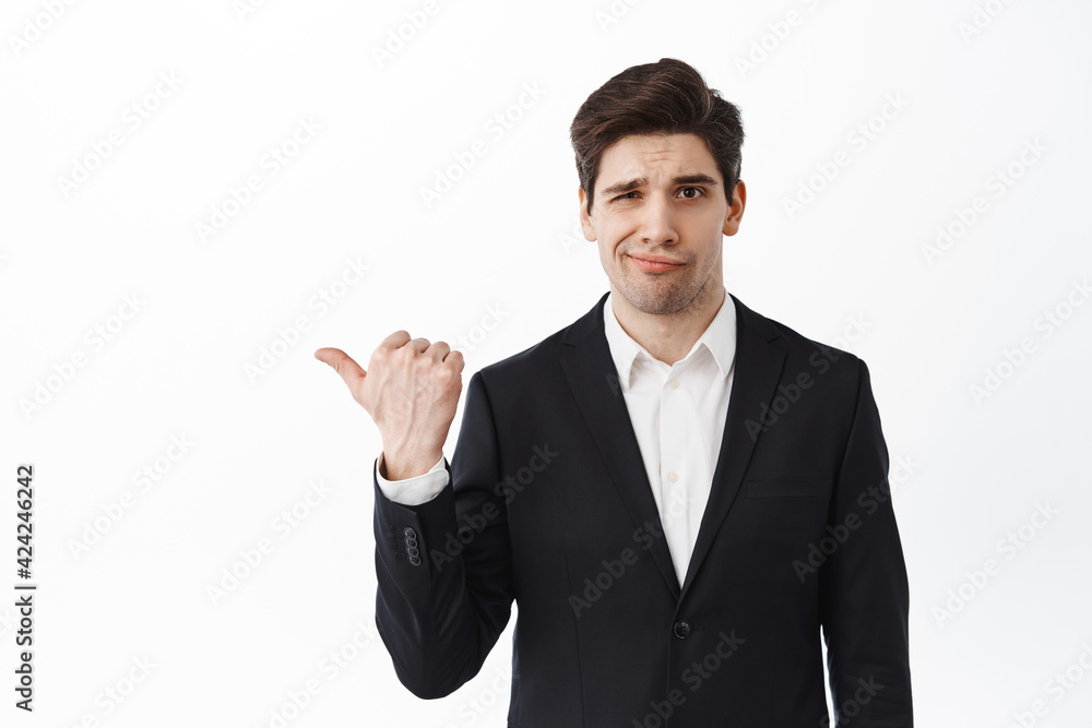 Disappointed business man frowning, pointing left at bad work, displeased or doubtful about advertisement, standing in suit against white background
