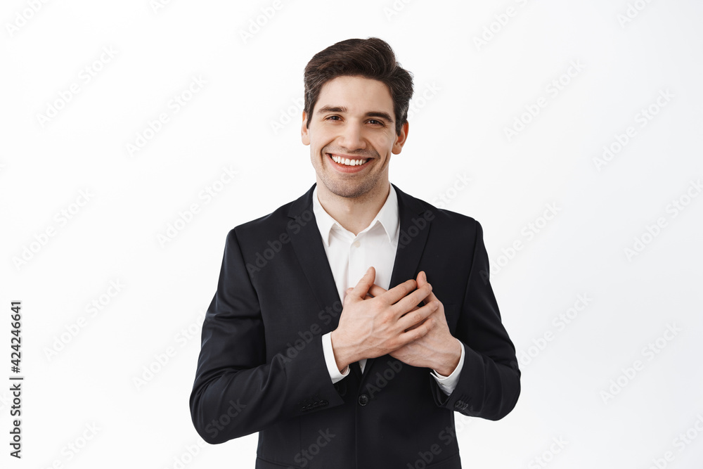 Company cares about clients. Smiling handsome business man in black suit hold hands on heart and looking caring and heartfelt at camera, standing over white background Company cares about clients. Smiling handsome business man in black suit hold hands on heart and looking caring and heartfelt at camera, standing over white background
