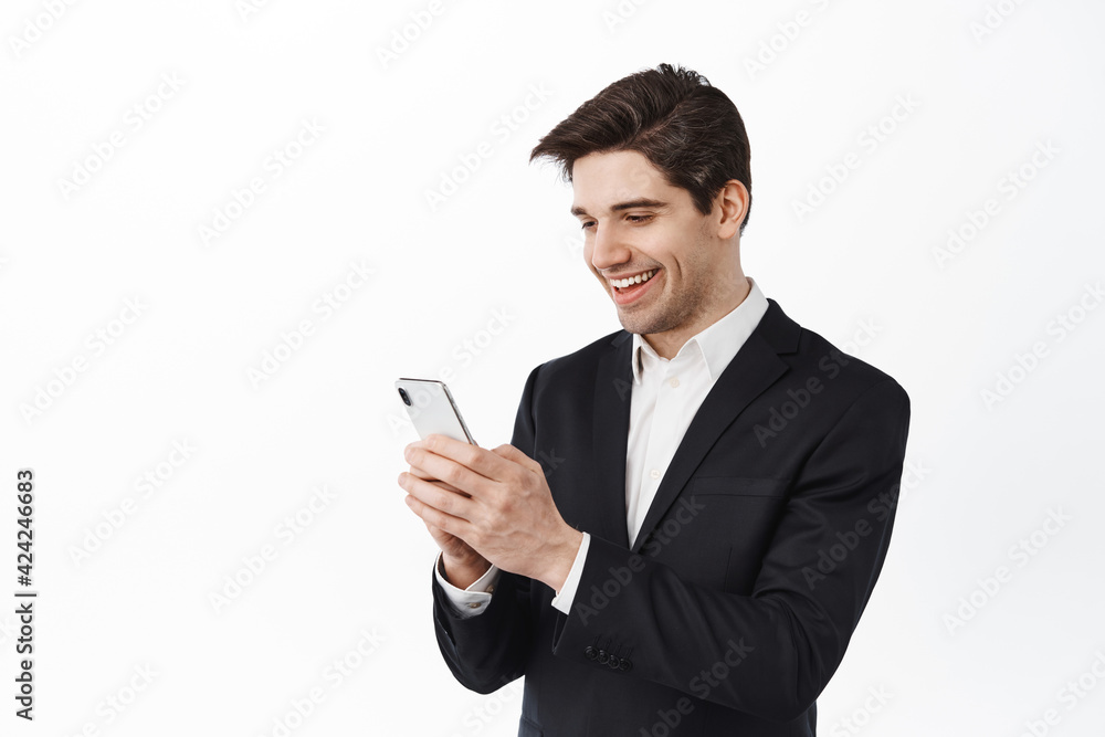 Handsome corporate man chatting on phone, texting message and looking at smartphone screen with pleased happy smile, standing over white background Handsome corporate man chatting on phone, texting message and looking at smartphone screen with pleased happy smile, standing over white background