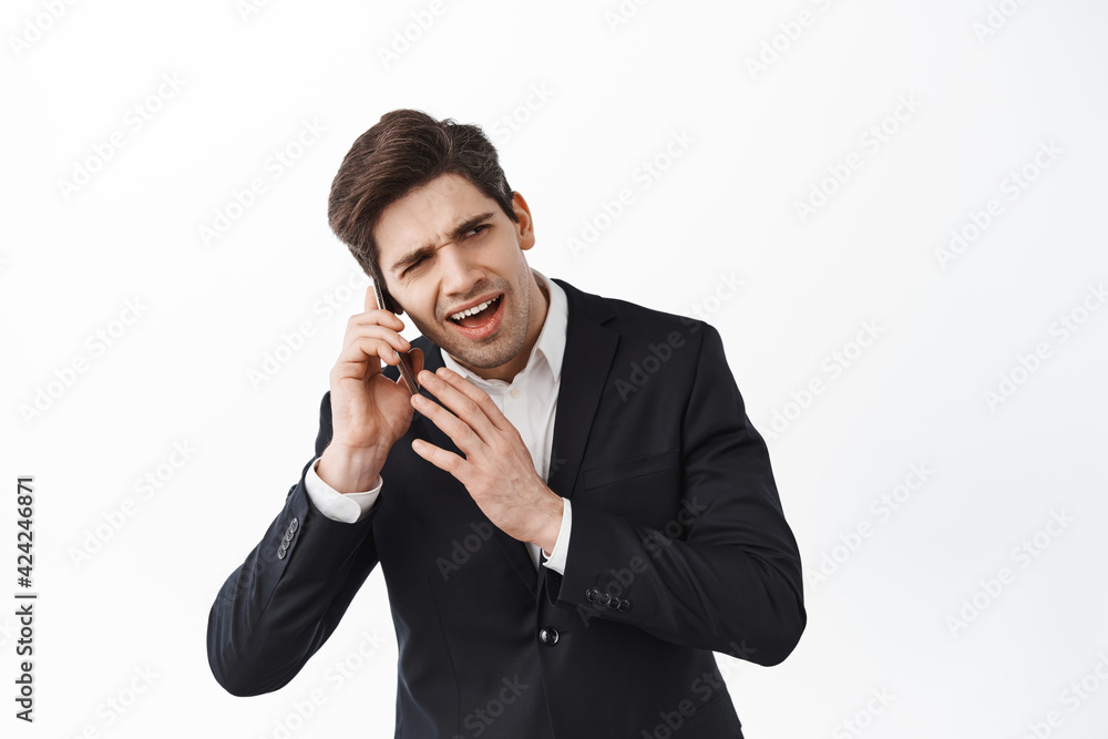 Business man cant hear person on the phone, having a call crowded loud place, frowning confused, standing in black suit against white background Business man cant hear person on the phone, having a call crowded loud place, frowning confused, standing in black suit against white background