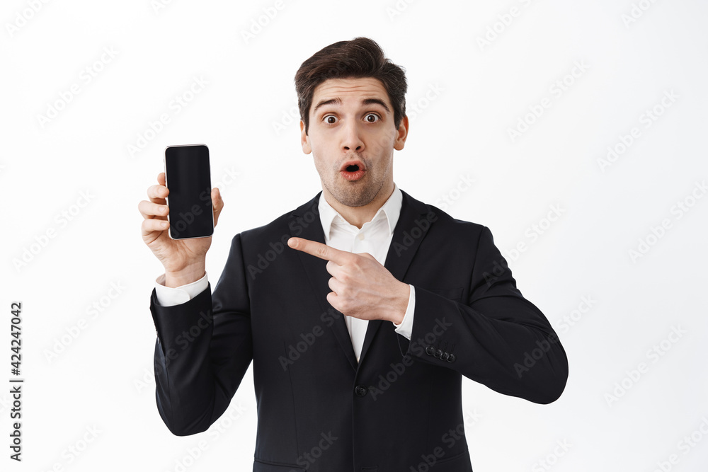 Impressed corporate man in suit pointing at smartphone screen, showing mobile website, standing over white background Impressed corporate man in suit pointing at smartphone screen, showing mobile website, standing over white background